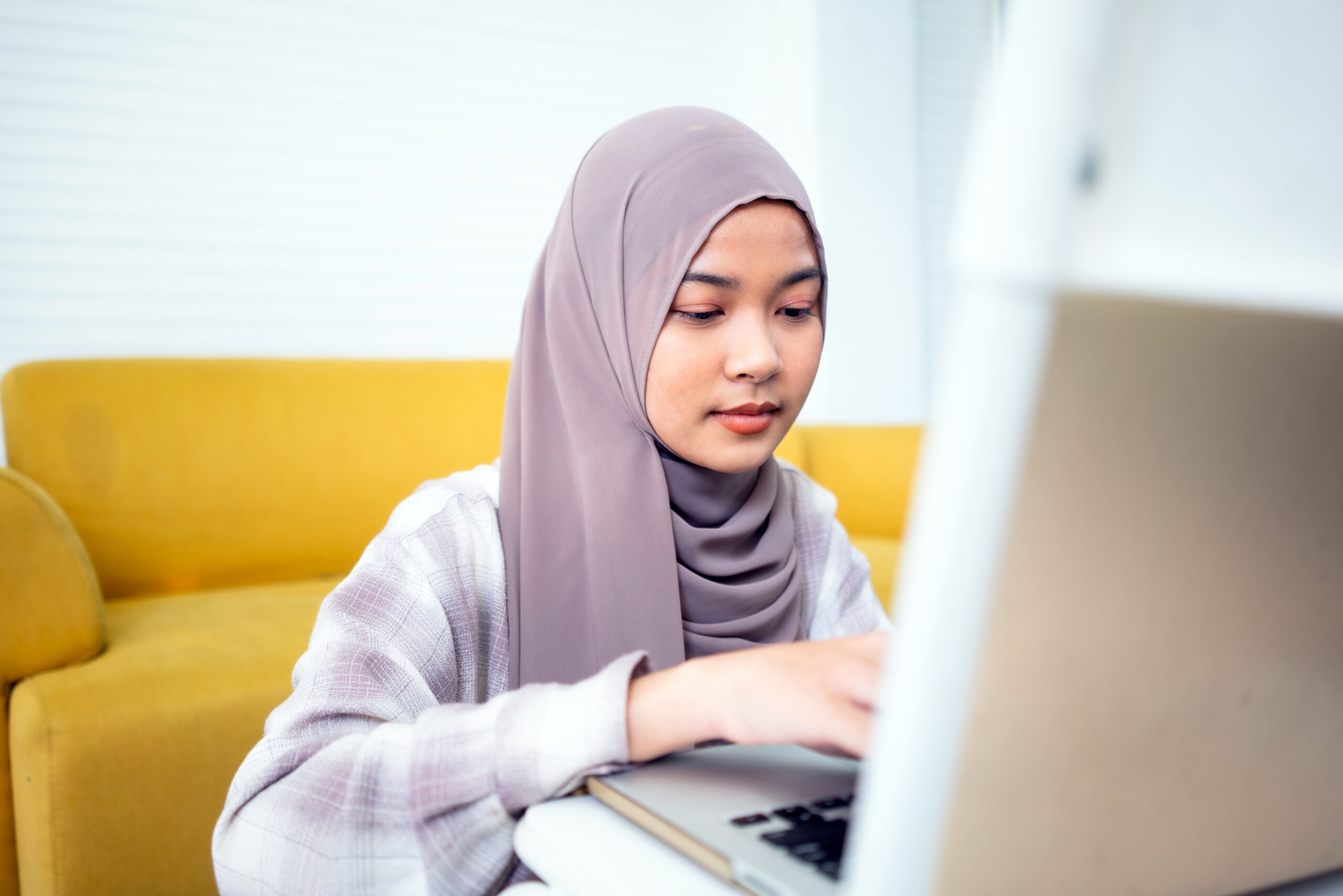 Young beautiful Muslim woman wearing hijab using laptop at home