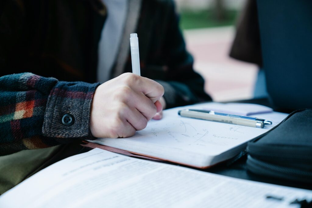 pexels-photo-7972309-7972309 Close-up of an individual writing in a notebook outdoors, featuring pens and paper.